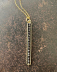 A gold-colored stick necklace with black diamond beads on a chain, displayed against a rustic background.