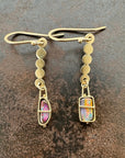 A pair of 18k gold dangle earrings with scalloped circular links and rectangular bezel-set iridescent boulder opal stones, displayed on a textured dark brown surface.