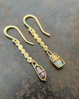 A pair of 18k gold dangle earrings with scalloped circular links and rectangular bezel-set iridescent boulder opal stones, displayed on a textured dark brown surface.