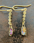 A pair of 18k gold dangle earrings with scalloped circular links and rectangular bezel-set iridescent boulder opal stones, displayed on a textured dark brown surface.