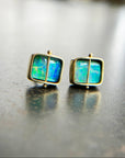 A pair of square stud earrings with iridescent blue-green boulder opal inlays in 18k gold settings, resting on a dark reflective surface.