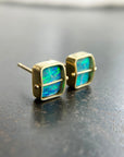 A pair of square stud earrings with iridescent blue-green boulder opal inlays in 18k gold settings, resting on a dark reflective surface.