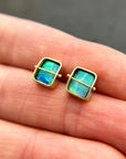 A pair of square stud earrings with iridescent blue-green boulder opal inlays in 18k gold settings, resting on a dark reflective surface.