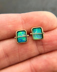 A pair of square stud earrings with iridescent blue-green boulder opal inlays in 18k gold settings, resting on a dark reflective surface.