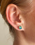 A pair of square stud earrings with iridescent blue-green boulder opal inlays in 18k gold settings, on a model.
