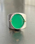 A wide silver band ring featuring a large, polished green chrysoprase teardrop-shaped gemstone in a ball rivet setting with small gold accents, resting on a textured gray surface.
