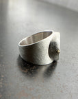 A wide silver band ring featuring a large, polished green chrysoprase teardrop-shaped gemstone in a ball rivet setting with small gold accents, resting on a textured gray surface.