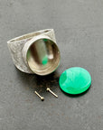 A wide silver band ring featuring a large, polished green chrysoprase teardrop-shaped gemstone in a ball rivet setting with small gold accents, resting on a textured gray surface.
