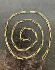 A gold elongated oval chain necklace coiled in a spiral on a textured gray surface.