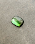 A polished green tourmaline cabochon with a cushion-cut shape, resting on a textured gray stone surface.
