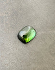 A polished green tourmaline cabochon with a cushion-cut shape, resting on a textured gray stone surface.