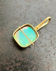 A square pendant with a gold-toned frame and loop, featuring a vibrant iridescent green-blue opal stone, resting on a textured dark gray surface.