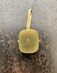 A gold pendant with a rounded rectangular cushion-cut face and a top loop, displayed on a textured dark gray and brown surface.