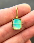 A gold pendant with a square iridescent opal stone held in a person's hand. The pendant features a gold bezel and loop, with a thin horizontal gold bar across the center.