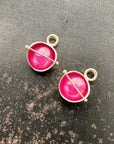 Two 14k gold hoop charms with vibrant pink ruby cabochon gemstones and a central gold bar, displayed on a dark textured surface.
