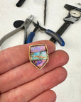 A shield-shaped pendant featuring an 18k gold frame and three rectangular inlay wood replacement opals in pink, turquoise, purple, and brown.