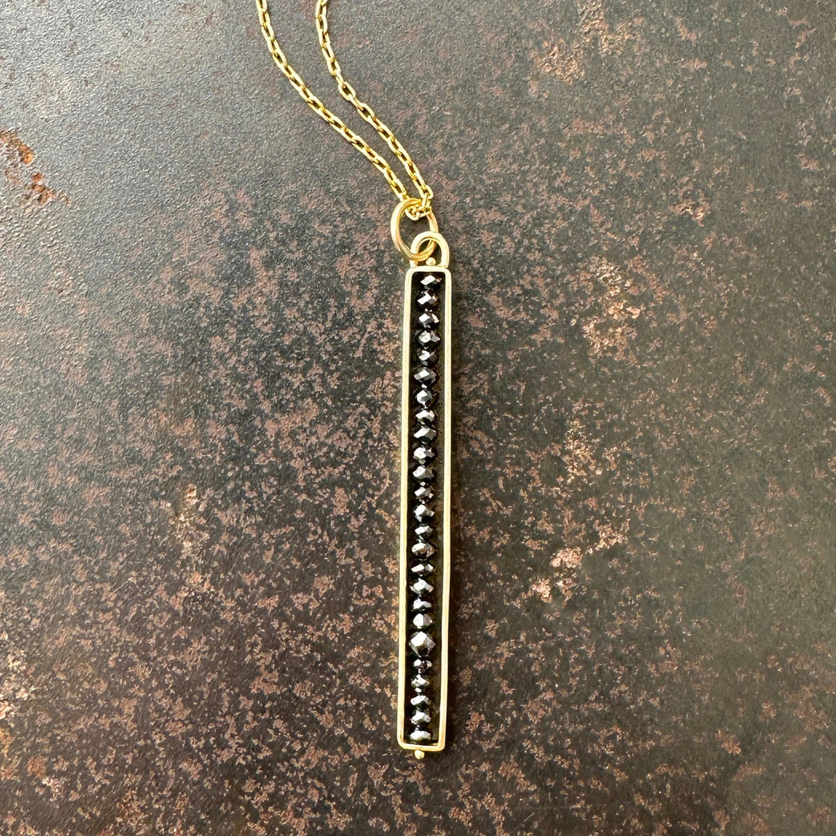 A gold-colored stick necklace with black diamond beads on a chain, displayed against a rustic background.
