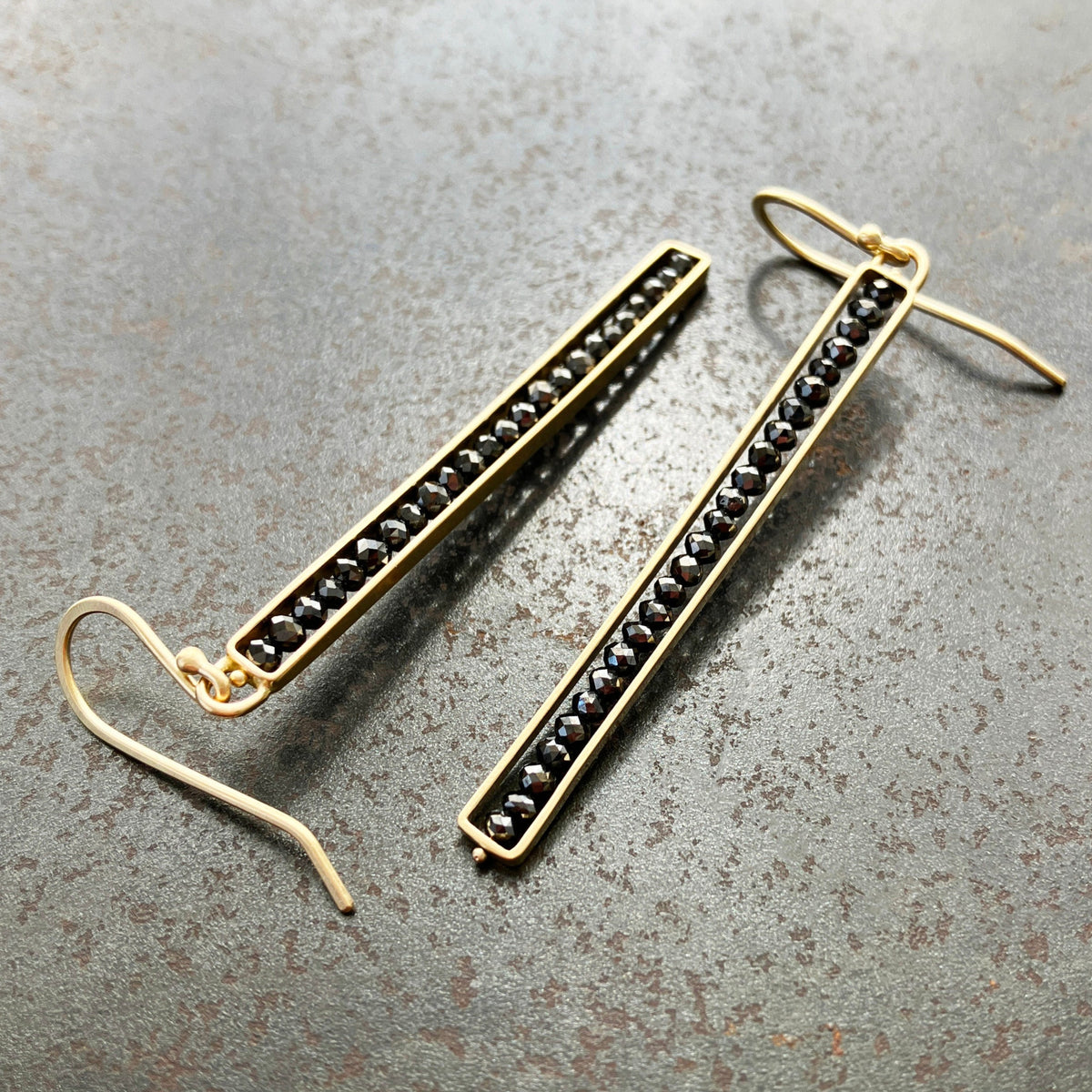 This image displays a pair of sophisticated Black Diamond Beaded Stick Earrings. The earrings feature long, slender 14k gold-frames, each adorned with a series of small black beads.