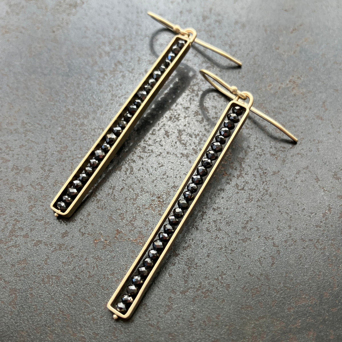 A pair of sleek stick earrings with black diamonds embedded in a line, set in 14k gold settings, displayed against a textured dark background.