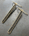 A pair of sleek stick earrings with black diamonds embedded in a line, set in 14k gold settings, displayed against a textured dark background.