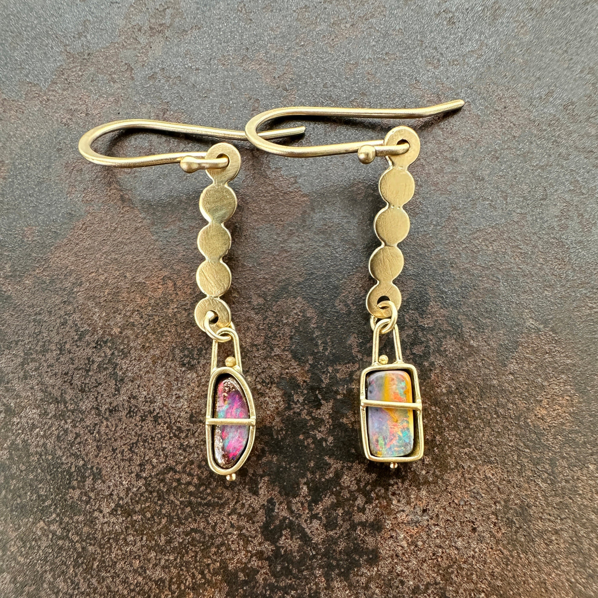 A pair of 18k gold dangle earrings with scalloped circular links and rectangular bezel-set iridescent boulder opal stones, displayed on a textured dark brown surface.