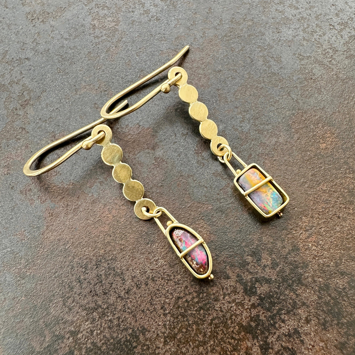 A pair of 18k gold dangle earrings with scalloped circular links and rectangular bezel-set iridescent boulder opal stones, displayed on a textured dark brown surface.