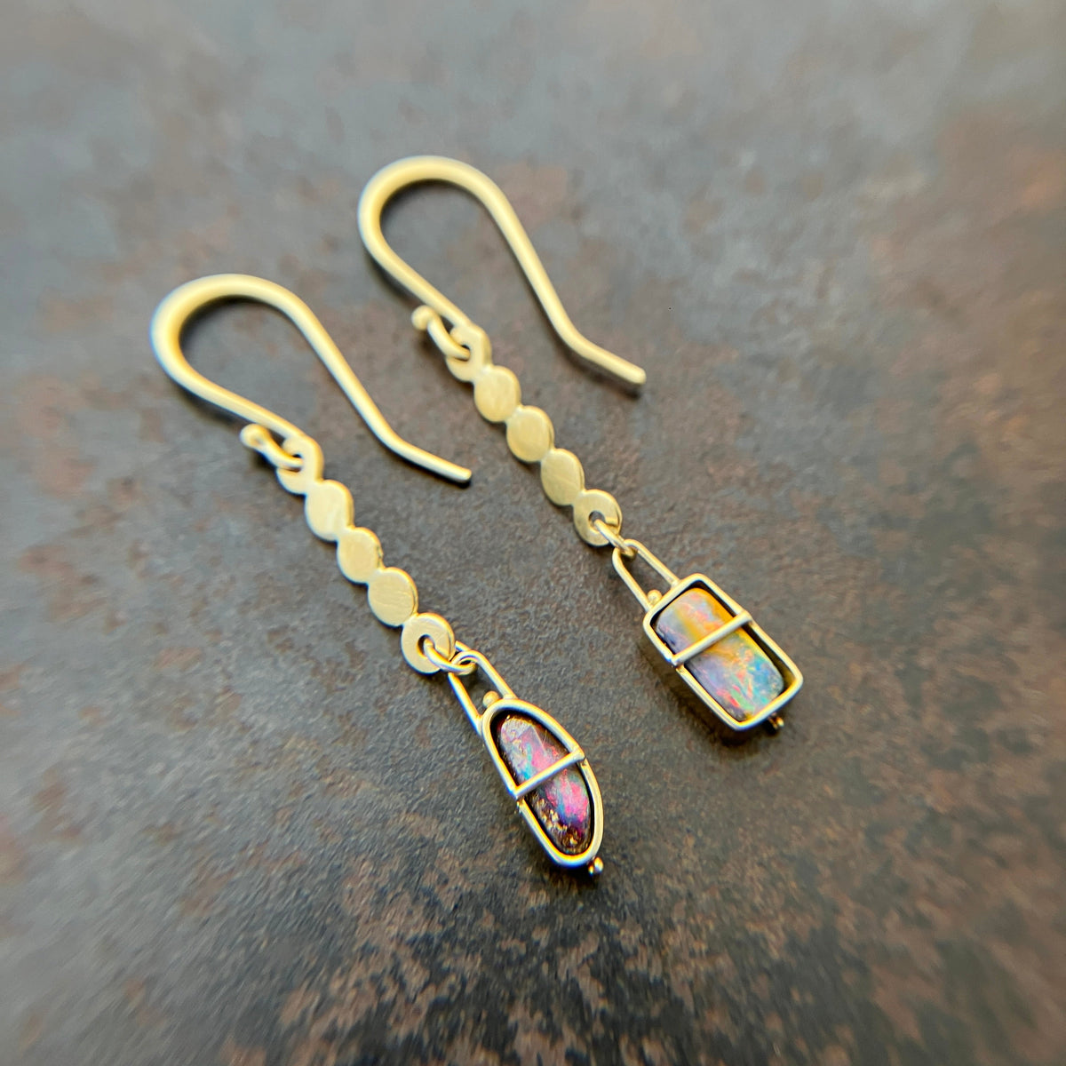 A pair of 18k gold dangle earrings with scalloped circular links and rectangular bezel-set iridescent boulder opal stones, displayed on a textured dark brown surface.