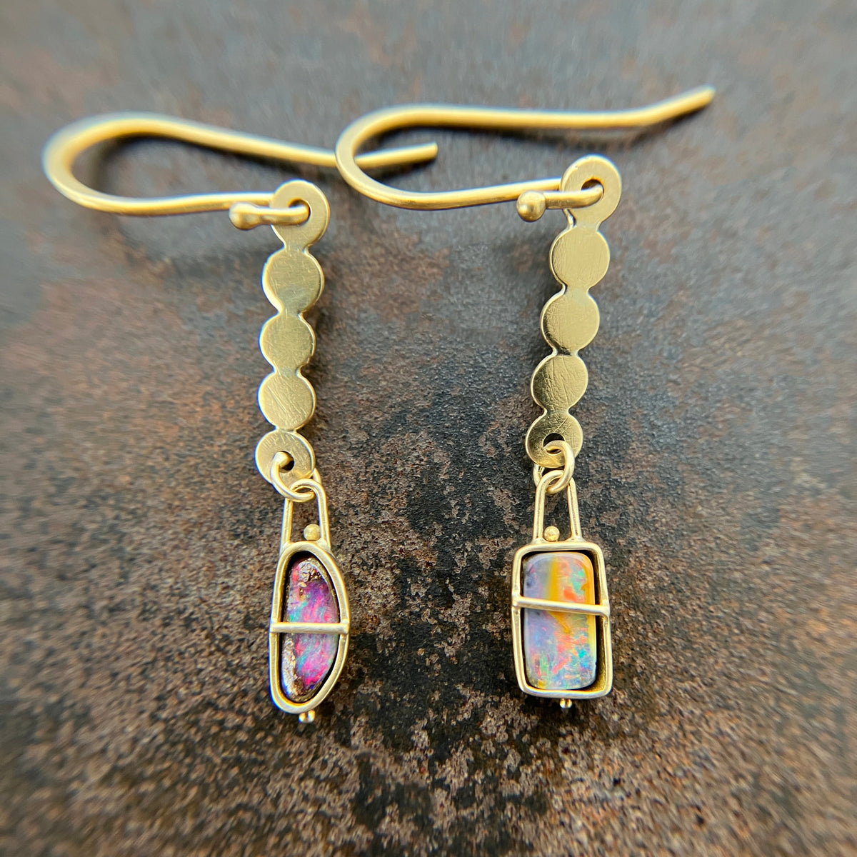 A pair of 18k gold dangle earrings with scalloped circular links and rectangular bezel-set iridescent boulder opal stones, displayed on a textured dark brown surface.
