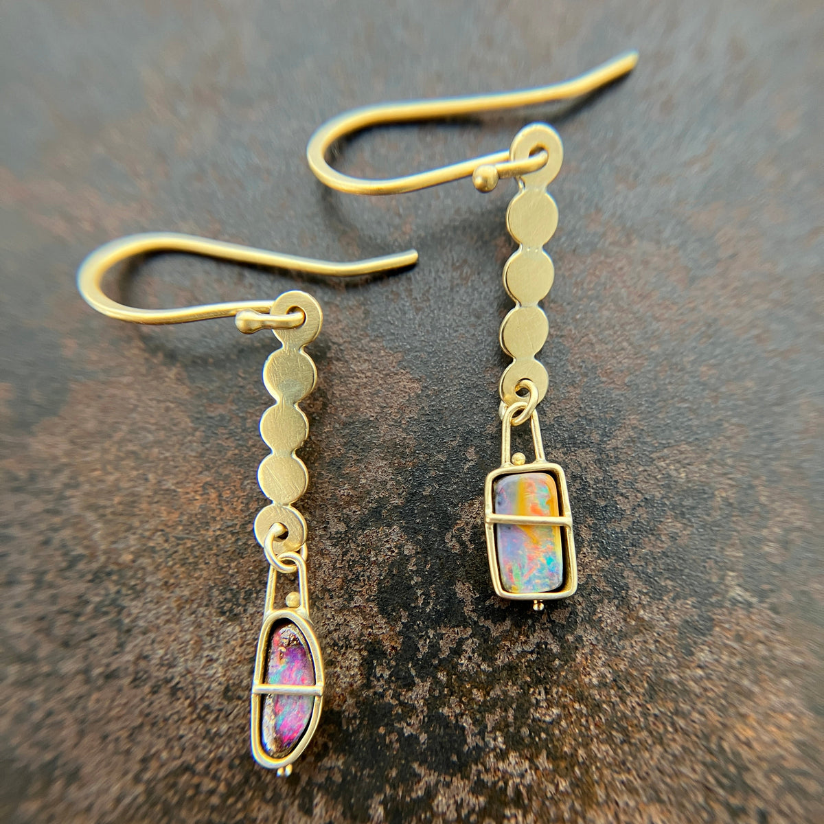 A pair of 18k gold dangle earrings with scalloped circular links and rectangular bezel-set iridescent boulder opal stones, displayed on a textured dark brown surface.