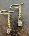 A pair of 18k gold dangle earrings with scalloped circular links and rectangular bezel-set iridescent boulder opal stones, displayed on a textured dark brown surface.