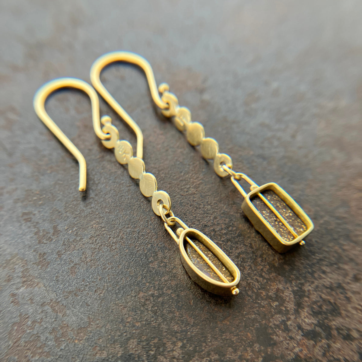 A pair of 18k gold dangle earrings with scalloped circular links and rectangular bezel-set iridescent boulder opal stones, displayed on a textured dark brown surface.