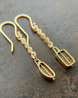 A pair of 18k gold dangle earrings with scalloped circular links and rectangular bezel-set iridescent boulder opal stones, displayed on a textured dark brown surface.