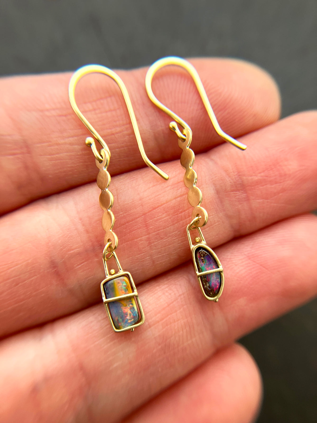 A pair of 18k gold dangle earrings with scalloped circular links and rectangular bezel-set iridescent boulder opal stones, displayed on a hand.