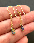 A pair of 18k gold dangle earrings with scalloped circular links and rectangular bezel-set iridescent boulder opal stones, displayed on a hand.
