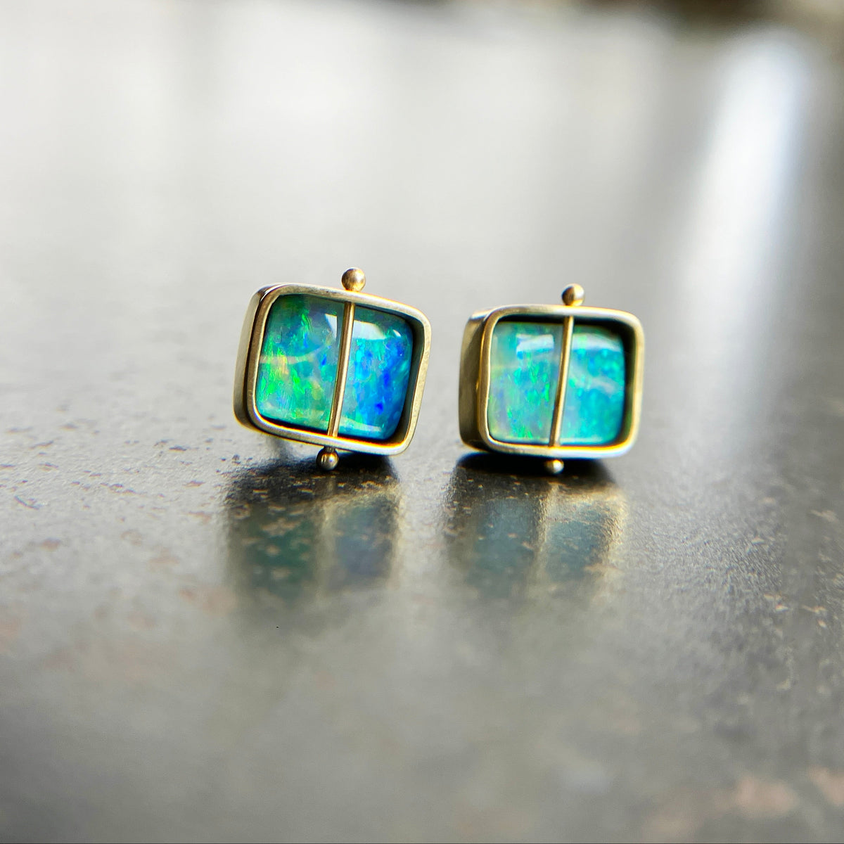 A pair of square stud earrings with iridescent blue-green boulder opal inlays in 18k gold settings, resting on a dark reflective surface.