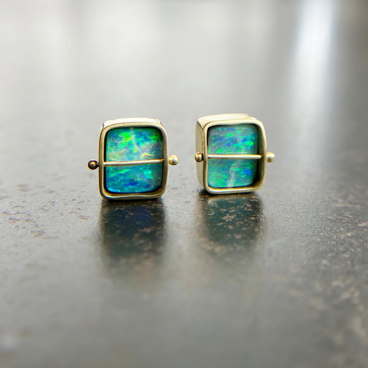 A pair of square stud earrings with iridescent blue-green boulder opal inlays in 18k gold settings, resting on a dark reflective surface.