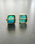 A pair of square stud earrings with iridescent blue-green boulder opal inlays in 18k gold settings, resting on a dark reflective surface.