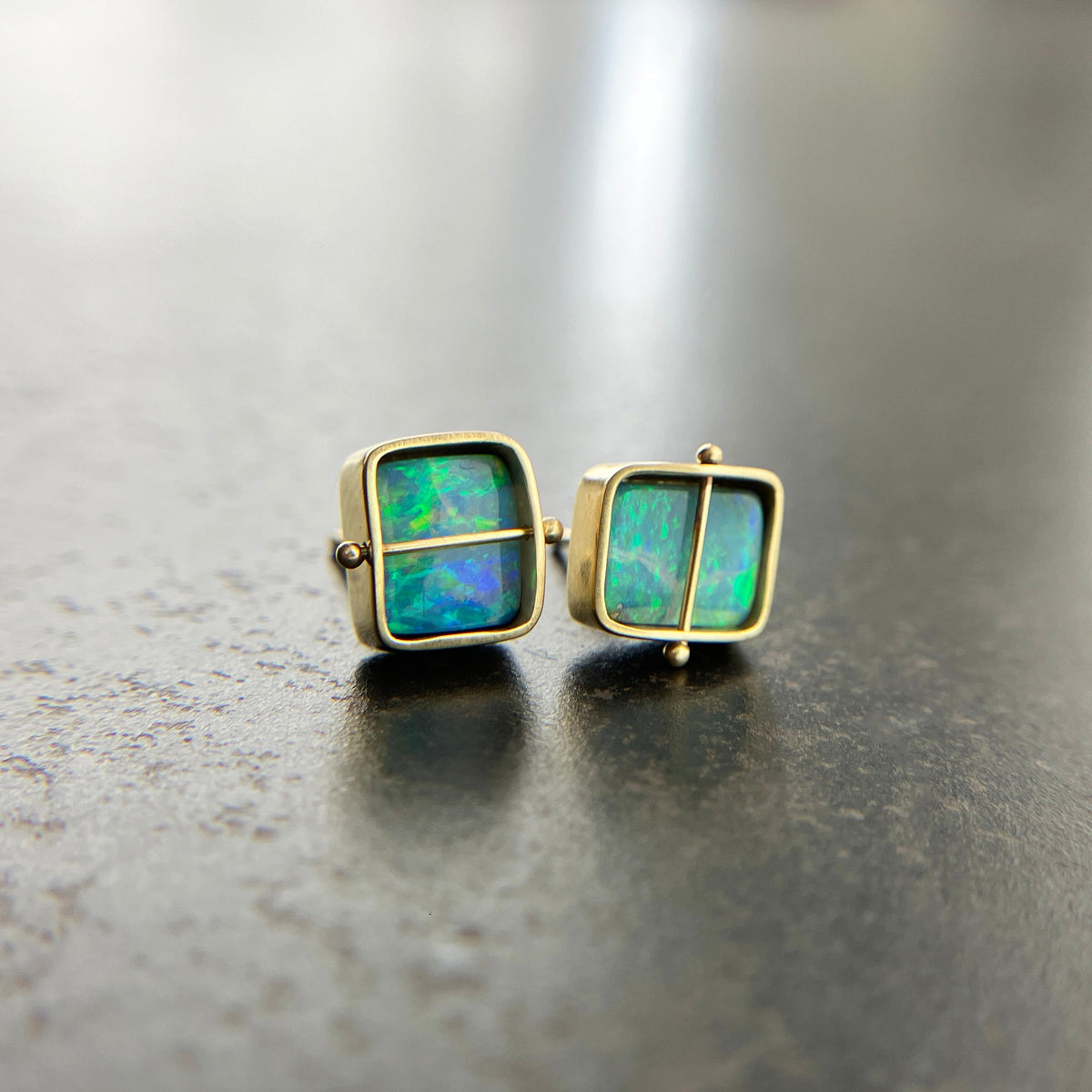 A pair of square stud earrings with iridescent blue-green boulder opal inlays in 18k gold settings, resting on a dark reflective surface.