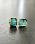 A pair of square stud earrings with iridescent blue-green boulder opal inlays in 18k gold settings, resting on a dark reflective surface.
