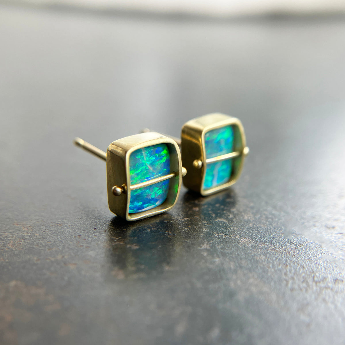 A pair of square stud earrings with iridescent blue-green boulder opal inlays in 18k gold settings, resting on a dark reflective surface.