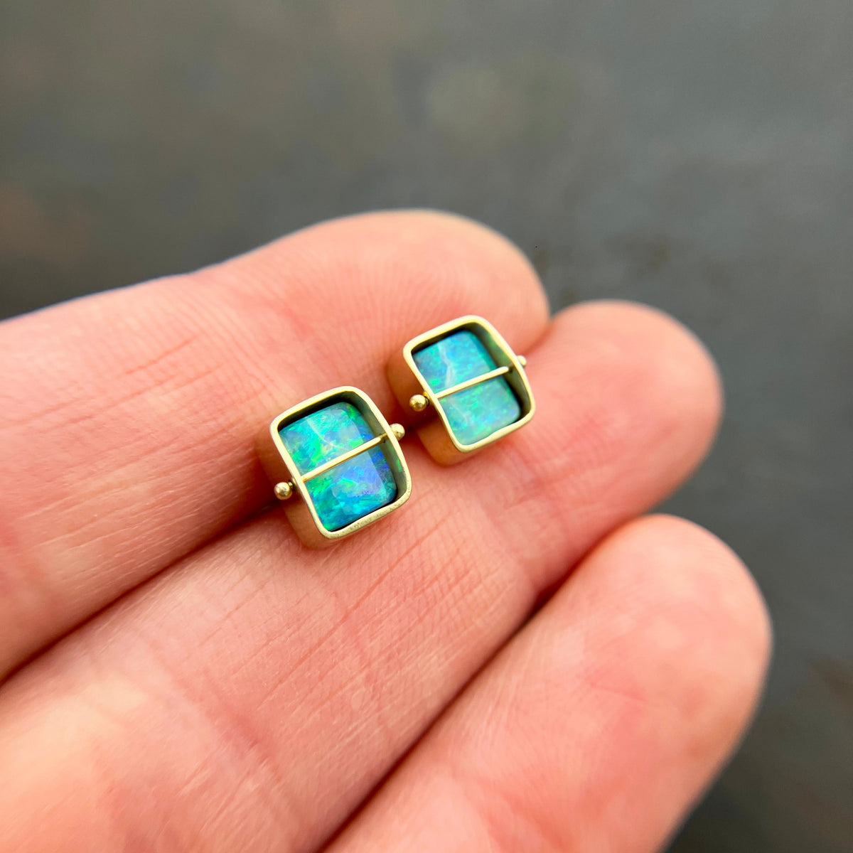 A pair of square stud earrings with iridescent blue-green boulder opal inlays in 18k gold settings, resting on a dark reflective surface.