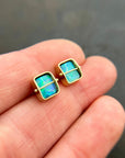 A pair of square stud earrings with iridescent blue-green boulder opal inlays in 18k gold settings, resting on a dark reflective surface.