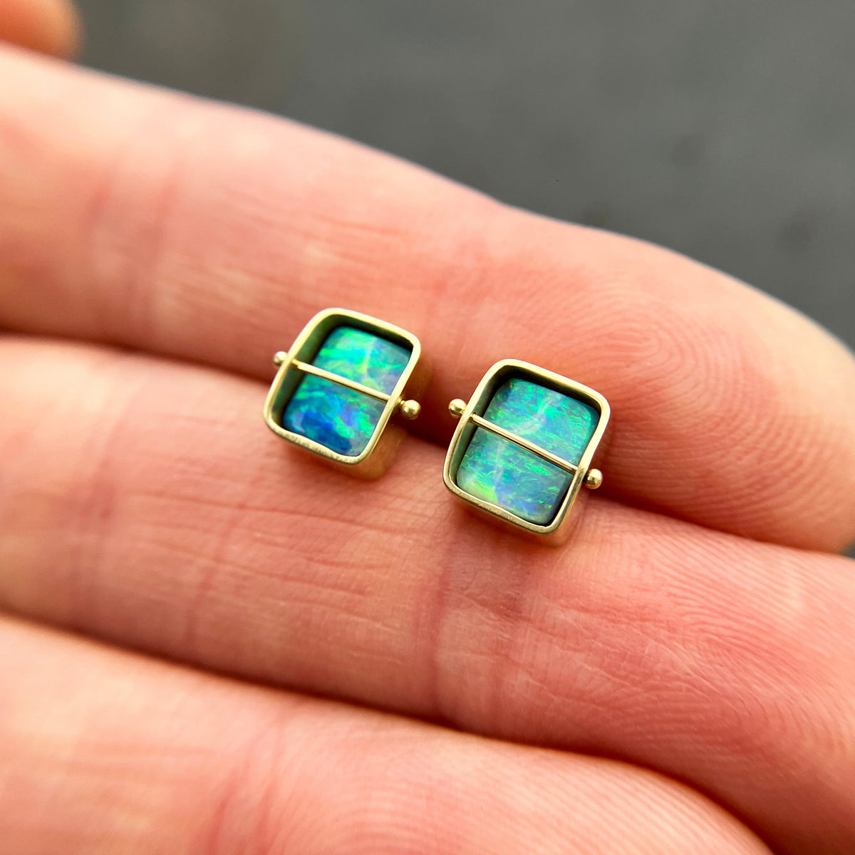 A pair of square stud earrings with iridescent blue-green boulder opal inlays in 18k gold settings, resting on a dark reflective surface.