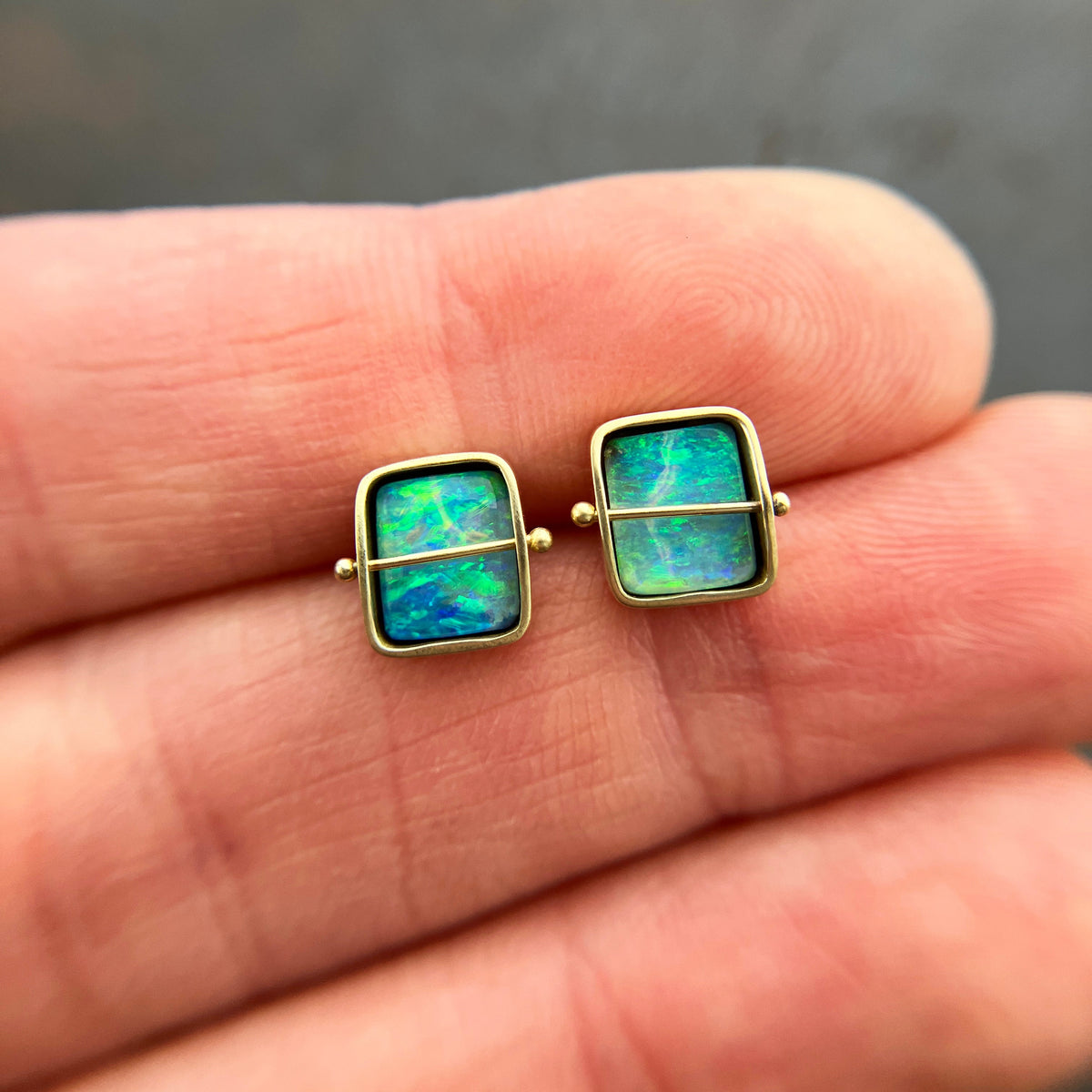 A pair of square stud earrings with iridescent blue-green boulder opal inlays in 18k gold settings, resting on a dark reflective surface.