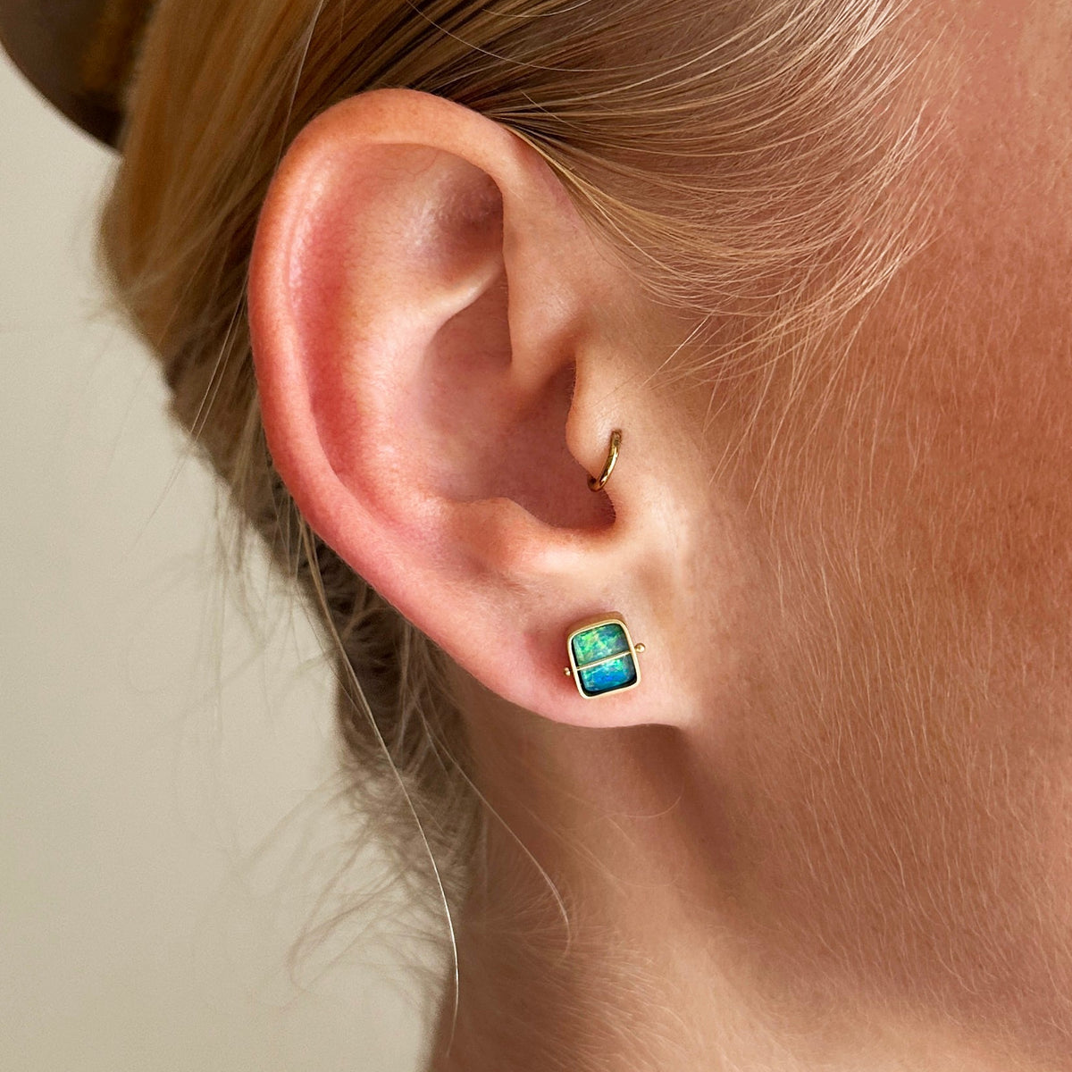 A pair of square stud earrings with iridescent blue-green boulder opal inlays in 18k gold settings, on a model.