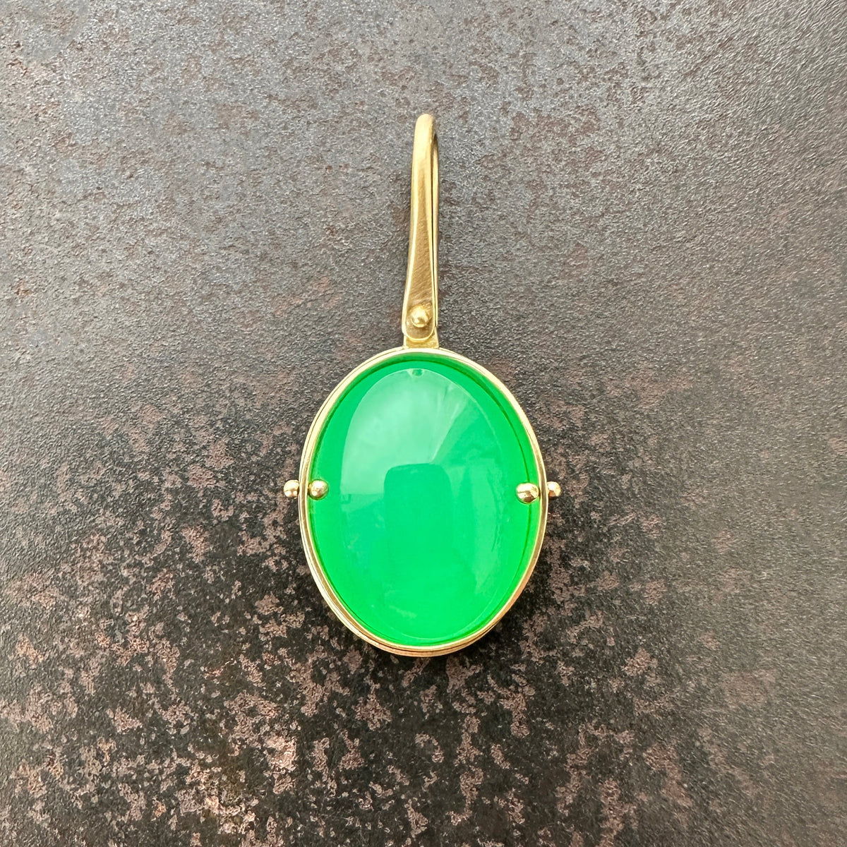 An oval pendant featuring a vibrant green chrysoprase gemstone in a gold bezel setting with a gold hook, displayed on a textured dark gray background.