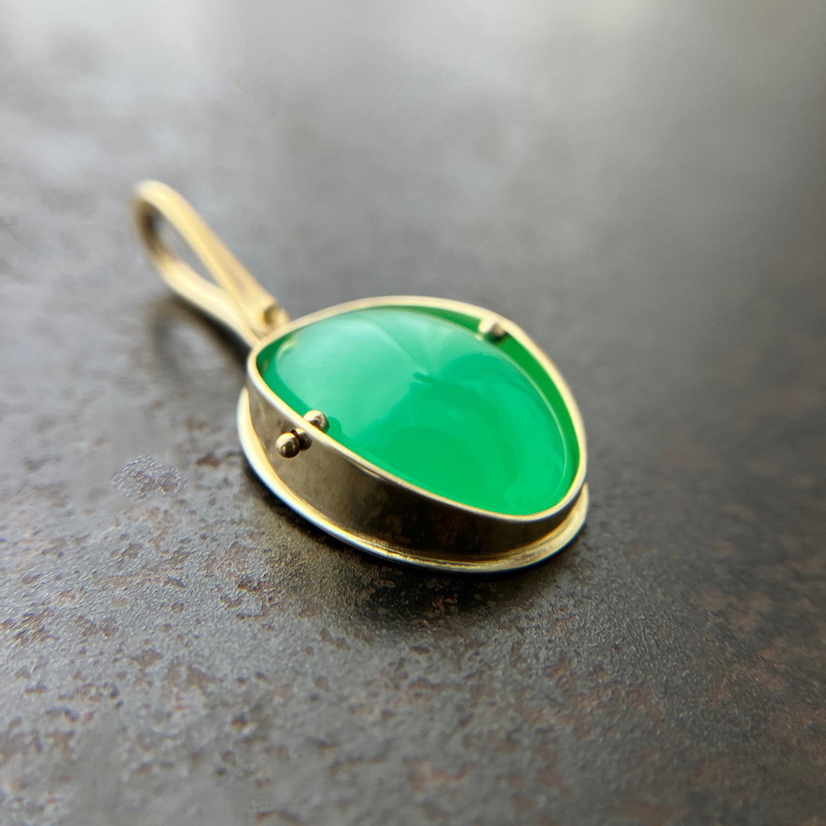 An oval pendant featuring a vibrant green chrysoprase gemstone in a gold bezel setting with a gold hook, displayed on a textured dark gray background.