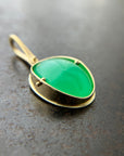 An oval pendant featuring a vibrant green chrysoprase gemstone in a gold bezel setting with a gold hook, displayed on a textured dark gray background.