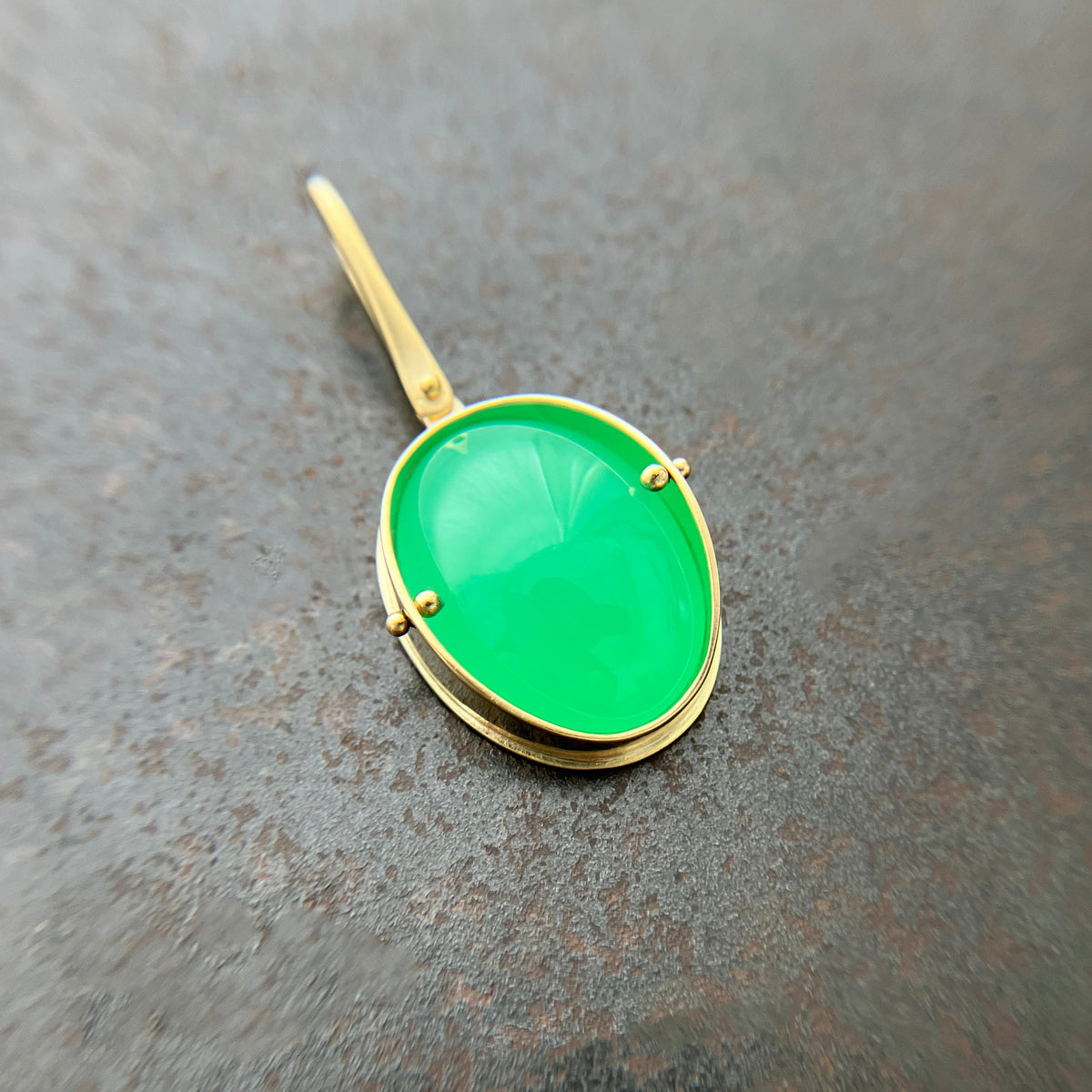 An oval pendant featuring a vibrant green chrysoprase gemstone in a gold bezel setting with a gold hook, displayed on a textured dark gray background.