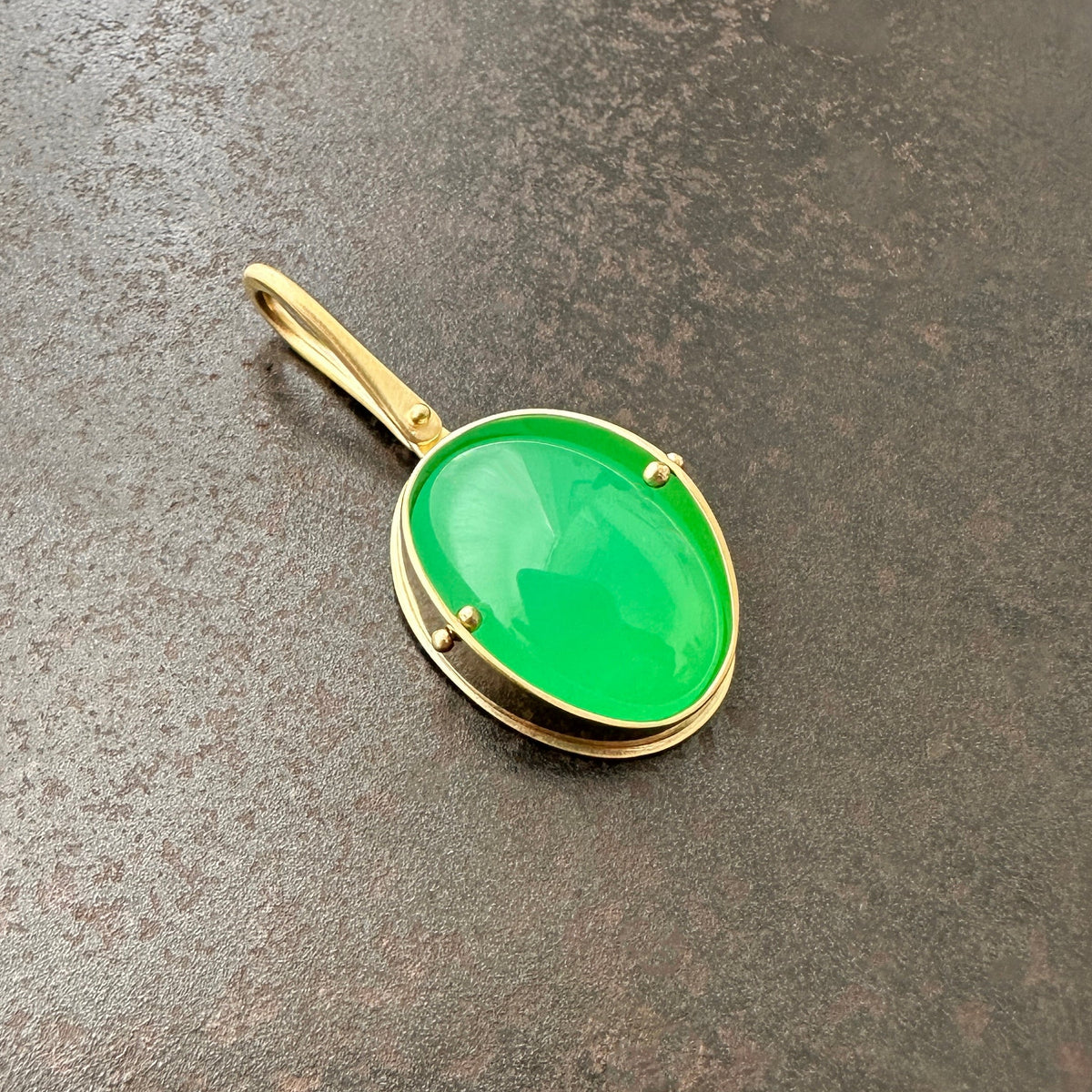 An oval pendant featuring a vibrant green chrysoprase gemstone in a gold bezel setting with a gold hook, displayed on a textured dark gray background.